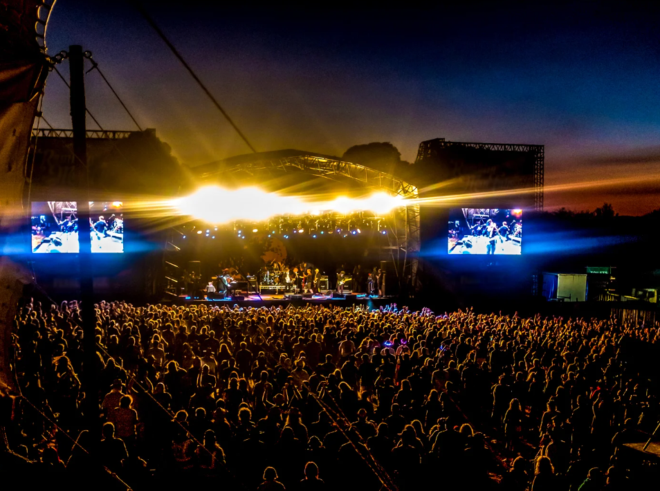 A large outdoor concert audience watching a brightly lit stage at dusk